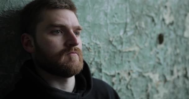 Close-up to the face of a sad man with a red beard who is feathers against an old cracked wall — Stock Video