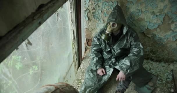 Strefa zagrożenia. Widok z góry. Mężczyzna w chemicznie ochronnej odzieży siedzi w pół zniszczonym pokoju przy oknie i patrzy przez okno. obok niego jest kanister. — Wideo stockowe
