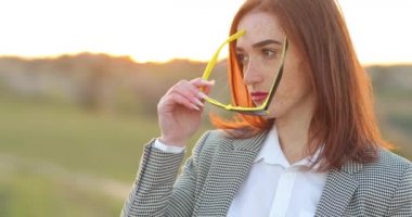 Açık havada, çilli, güzel, modaya uygun, kızıl saçlı bir kızın portresi. Model siyah giyiyor, sarı gözlükler takıyor, şık kıyafetler giyiyor, yan bakıyor. Gün batımı