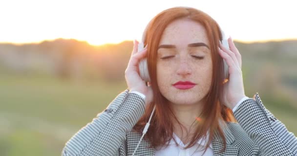 Wanita muda berambut merah wajah dengan bintik-bintik. Gadis berambut merah yang menarik mendengarkan musik di headphone dengan mata tertutup. — Stok Video