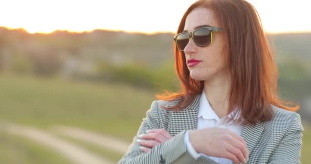 Porträt einer stylischen attraktiven rothaarigen jungen Frau mit Sommersprossen im Gesicht und Sonnenbrille steht in der Natur und blickt geradeaus in die Kamera. Junge Frauen nimmt ihre Brille — Stockvideo