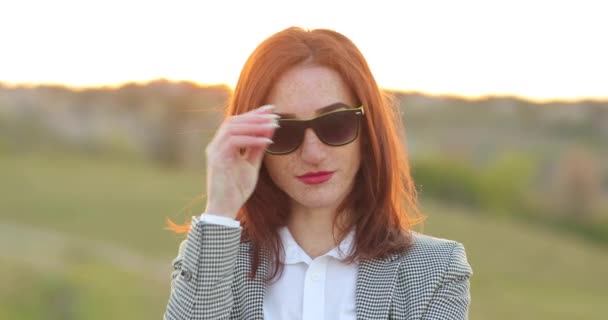 Πορτρέτο της ελκυστική κομψή κοκκινομάλλα επιχειρηματίας με πολλές φακίδες στο πρόσωπό της κοιτάζοντας κατ 'ευθείαν στην κάμερα και την απογείωση γυαλιά ηλίου. — Αρχείο Βίντεο