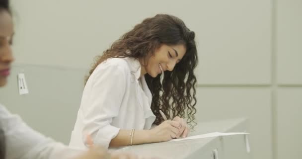 Atrakcyjna hinduska dziewczyna z długimi kręconymi włosami siedzi na wykładzie na uniwersytecie medycznym i pisze. — Wideo stockowe