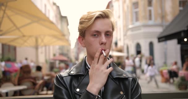 Portrait d'un jeune homme séduisant debout dans la rue et fumant une cigarette. derrière lui les gens marchent et s'assoient dans un café — Video