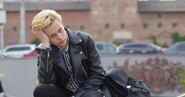 Sad attractive blond guy sitting on a bench in the city and resting his head on his arm. behind the castle and drive cars — Stock Video