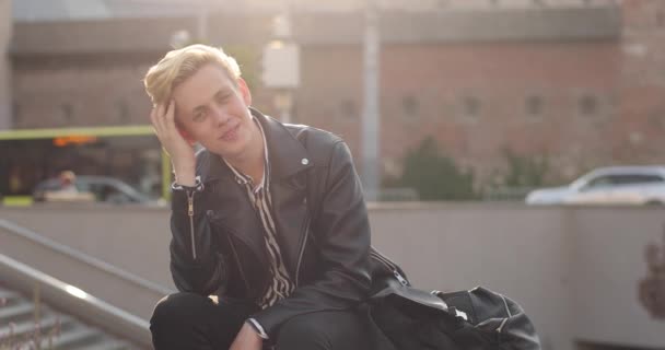 Привлекательный блондин сидит в городе на скамейке и бежит рукой по волосам. — стоковое видео