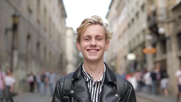 Ein gut gelaunter attraktiver junger Mann in schwarzer Jacke steht auf der Straße und lächelt. blond — Stockvideo
