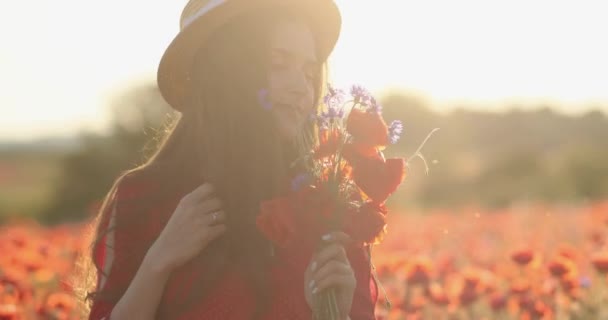 Mulher sorridente no meio do campo de papoilas a arrefecer ao pôr do sol. Jovem senhora morena em um vestido de bolinhas vermelhas e chapéu de palha segurando buquê de papoilas nas mãos. Dia de verão e atmosfera calma. 4K — Vídeo de Stock