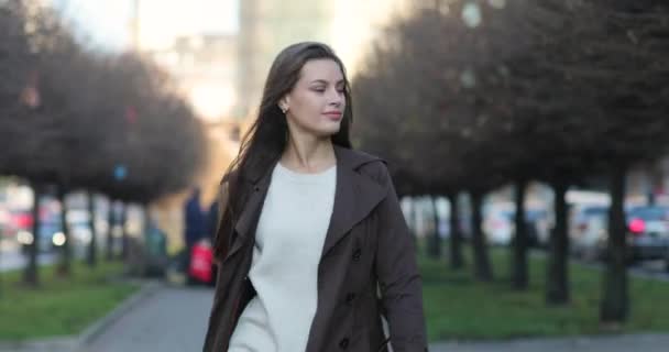 Femme confiante au regard fascinant en manteau marron marche sur une ruelle et joue avec ses cheveux par temps ensoleillé. Paramètres urbains en arrière-plan — Video