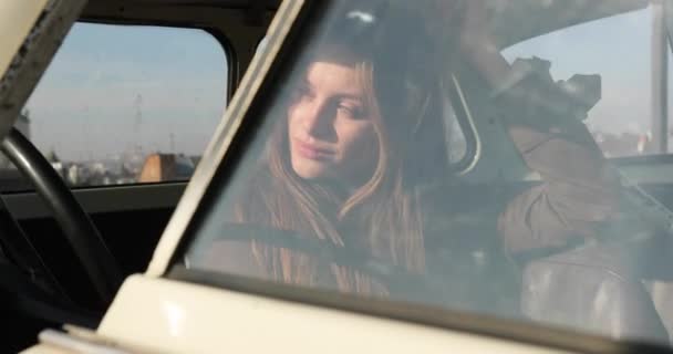 Stylish brunette woman is sitting alone in the white car on the roof while rays of sun shines on her. On the background panorama of old city in sunny day — Stock Video