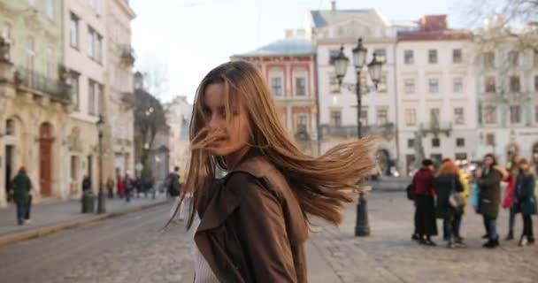 Sparando da dietro di donna attraente in cappotto marrone, sta camminando sulla piazza. Si gira e guarda in macchina fotografica mentre i suoi capelli lunghi bruna soffia nel vento nella giornata di sole. Vecchi edifici belli — Video Stock