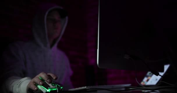 Atrakcyjny młody mężczyzna w czapce i kapturze pracujący przy komputerze w ciemnym pokoju z różowym oświetleniem. Facet kontroluje mysz z zielonymi i fioletowymi światłami. komputer, klawiatura i mysz są na stole — Wideo stockowe