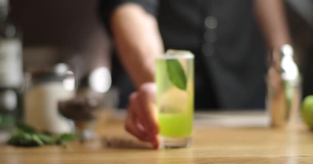 Крупным планом крепкий бармен протягивает коктейль. зеленый коктейль с лавровым листом на столе — стоковое видео