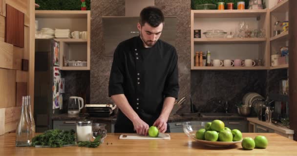 Atraktivní mladý kuchař v černé konvice v kuchyni krájí zelená jablka. spousta koření na stole před ním. — Stock video