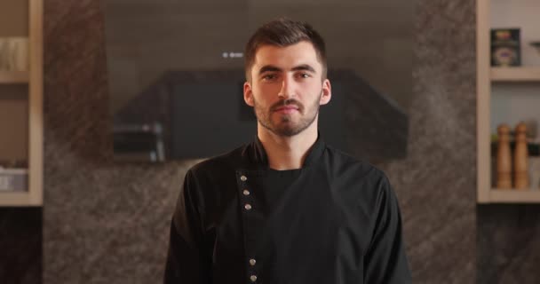Πορτρέτο του σεφ σε μαύρο βραστήρα στέκεται στην κουζίνα και κοιτάζοντας κατ 'ευθείαν στην κάμερα. πίσω του είναι μια κουκούλα καθρέπτης κουζίνας, σκεύη και μπαχαρικά. πορτρέτο του ελκυστικό σεφ με γενειάδα σε μαύρο βραστήρα — Αρχείο Βίντεο