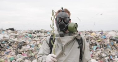 Gaz maskeli bir adam çöplerin arasında duruyor ve elinde tomurcukları olan bir dal tutuyor. Ekoloji. Felaket. Halkın sorunu. Atık doldurma sorunu.