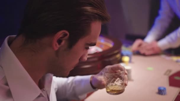 Hombre morena agitado en camisa blanca beber whisky mientras está sentado solo detrás de la mesa de ruleta en el Casino — Vídeo de stock
