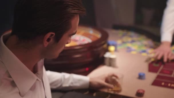 Hombre morena agitado en camisa blanca beber whisky mientras está sentado solo detrás de la mesa de ruleta en el Casino. Hombre enojado después de perder en el casino — Vídeos de Stock