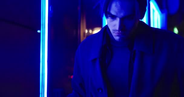 暗いコートと帽子に身を包んだブルネットの男が夜に通りを歩く。青い光の効果。夜市灯 — ストック動画