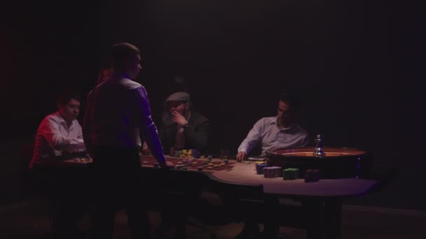 Eine Gruppe Männer sitzt hinter dem Roulettetisch im Casino, trinkt Whiskey und raucht Zigarren. Croupier im weißen Hemd dreht das Rad — Stockvideo
