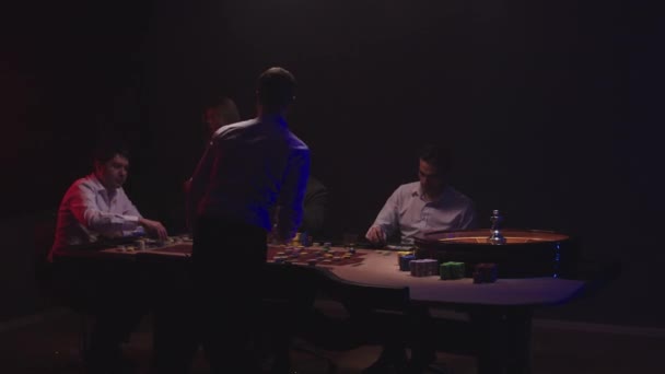 Група чоловіків сидить за таблицею Роулетт у казино, п'ючи віскі і куривши сигари. Крупір у білій сорочці крутить колесо. — стокове відео
