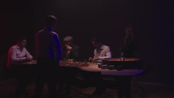 Группа мужчин сидит за столом рулетки в казино, пьет виски и курит сигары. Крупье в белой рубашке крутит колесо — стоковое видео