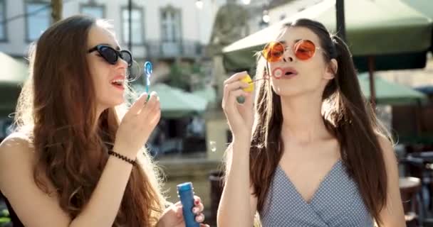 İki neşeli genç bayan dışarıda büyük sabun köpükleri ile oynuyorlar. Güneş gözlüklü iki güzel kız sabun köpüğü şişiriyor.. — Stok video