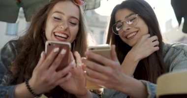 İki mutlu genç kadının iki akıllı telefon kullanışının ön görüntüsü. Açık havada, parkta, kampüste ya da kafede medyanın içeriğini izleyen iki güzel öğrenci.