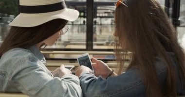İki kız arkadaş bankta oturup konuşuyor. İki çekici kız konuşarak ve akıllı telefon kullanarak eğleniyor. arka plan