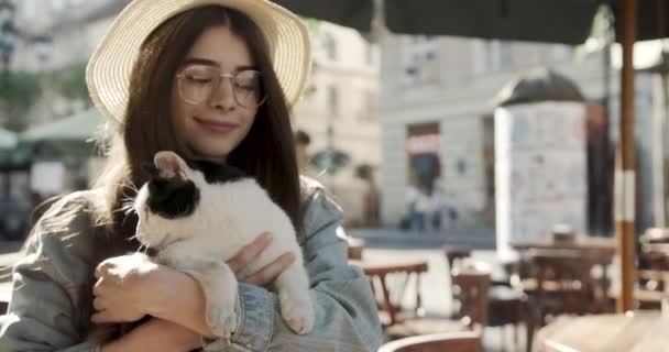 Chica de moda en una ciudad. Dama con gato. chica atractiva en un sombrero de paja con gafas fuertes con un gato en sus manos en la calle. — Vídeo de stock