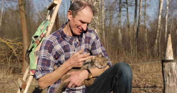 Un homme à la campagne est assis sous un arbre et joue avec un chat gris. senior en civil tenant un chat dans ses bras — Video