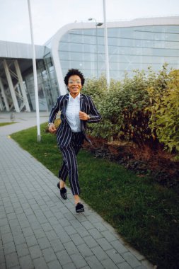 4 bin. Seyahat et, Dijital. Zarif çizgili takım elbiseli çekici bir Afrikalı Amerikalı kadın kameraya bakıyor, koşuyor, gülümsüyor ve kendini harika hissediyor. Havaalanının yakınındaki çalılıklara doğru koştu. İş kadını.