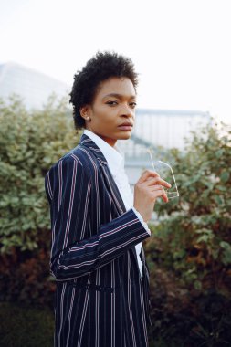 4 bin. Seyahat et, Dijital. Zarif çizgili takım elbiseli çekici bir Afrikalı Amerikalı kadın kameraya bakıyor, gözlüklerini tutuyor ve uçuşunu bekliyor. Arka plandaki çalıların üzerinde duruyor.