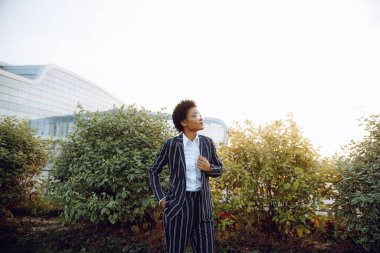 4 bin. Seyahat et, Dijital. Zarif çizgili takım elbiseli çekici bir Afrikalı Amerikalı kadın gökyüzüne bakıyor ve uçuşunu gülümseyerek bekliyor. Havaalanının yakınındaki çalıların arkasında duruyor.