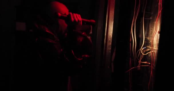 In een donkere kamer met een rood licht loopt een man met een zaklamp in een gasmasker en speciale kleding. elektriciteitsleidingen aan de muur — Stockvideo