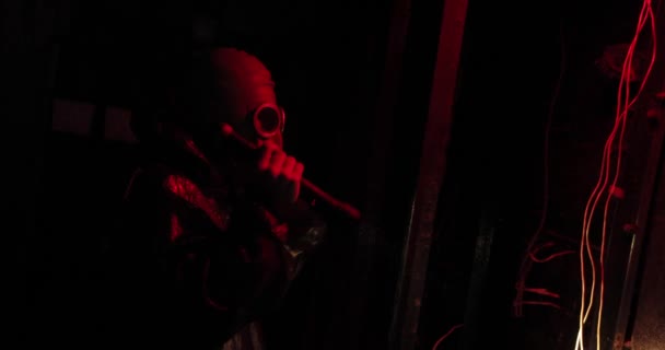 Lampu merah. Pria dengan pakaian khusus dan masker gas berdiri di ruangan gelap dan memegang senter di tangannya. seorang pria bersinar senter di dinding di mana menggantung kabel — Stok Video