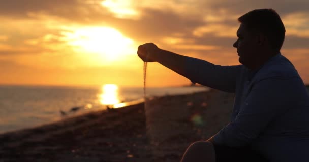 En mann sitter ved havet, og sanden faller av hans hånd. Ekstremt vakker solnedgang. – stockvideo