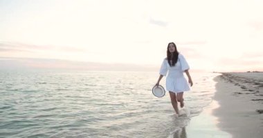 Güzel, otantik bir kadın gün batımında boş kumsalda sörf dalgalarıyla koşuyor. Kameraya gül ve gülümse, gerçek duygular ve duygular. Gelecek umuduyla tasasız bin yıllık gençlik..