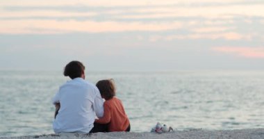 Baba, Kum Sahili kıyısında otururken Oğula Kucaklıyor. Aile Kavramı ve Doğada Birlikte Zaman Geçirmek. Baba ve Oğul Kum Sahili 'nde otururken Deniz Dalgalarına Bakar