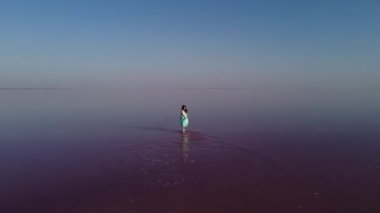 Güneşli bir yaz gününde pembe tuz gölünde mutlu bir kadın. Nane renginde elbisesi olan bir kadının pembe bir tuz gölünde yürüdüğü hava manzarası.
