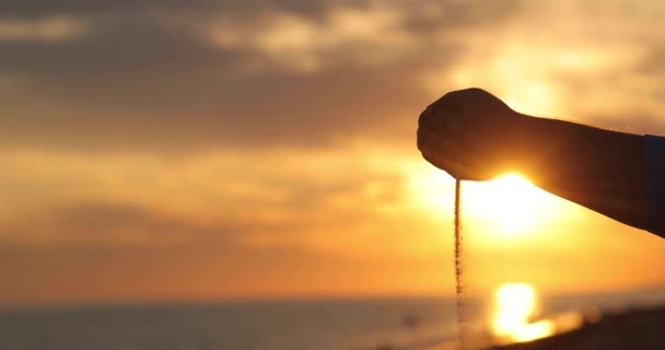 Man hand med sand häller på bakgrunden av en vacker solnedgång. — Stockvideo