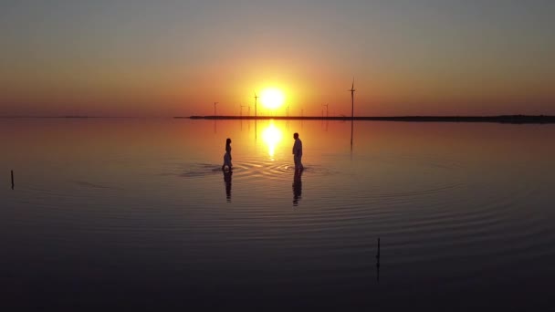 Homem e mulher vão se encontrar na água atrás do pôr-do-sol e moinhos de vento — Vídeo de Stock