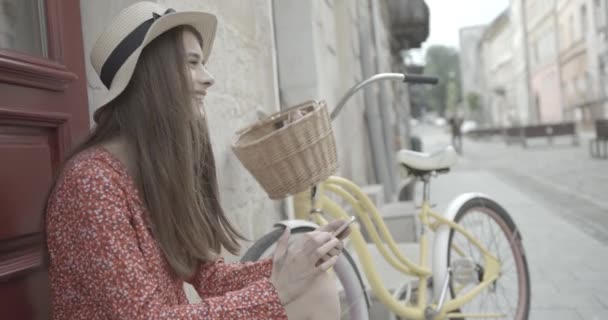 Молодая очаровательная женщина-велосипедистка с длинными темными волосами счастливо болтает, пишет смс и просматривает мобильный телефон, сидя на размытом фоне велосипеда. — стоковое видео