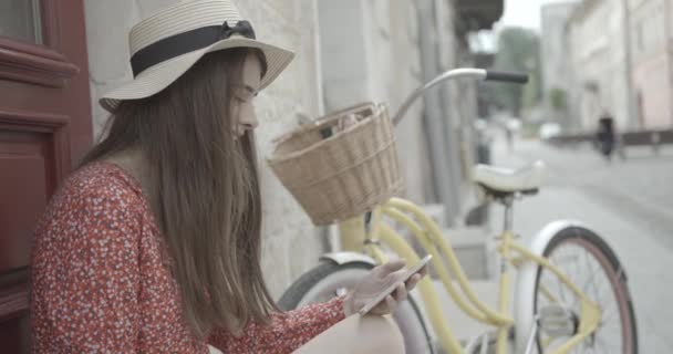 Portret pięknej brunetki odpoczywającej na świeżym powietrzu podczas rozmowy przez telefon komórkowy na rozmytym tle roweru. Młoda dziewczyna macha do kogoś. 4k. — Wideo stockowe