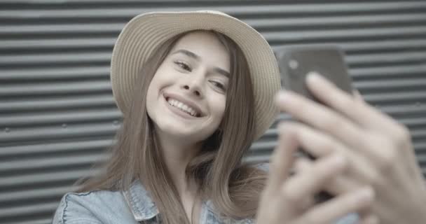 Positiva bruna sorridente in elegante cappello e giacca di jeans sta prendendo selfie sul telefono cellulare sullo sfondo isolato scuro. 4k. — Video Stock