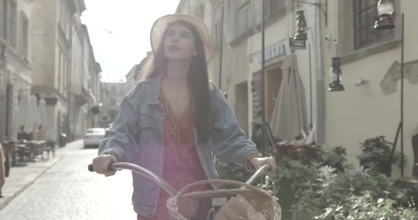 Giovane bella donna viaggiatore sta andando in bicicletta lungo le vecchie strade soleggiate. Indossa un vestito rosso, giacca e cappello di jeans. 4k filmati. — Video Stock