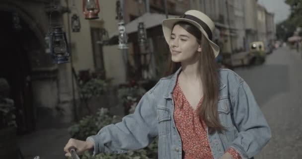 Bella ragazza felice con i capelli scuri sta camminando in bicicletta lungo le affascinanti strade durante il tramonto. Attraente turista indossa abito, giacca di jeans e cappello. — Video Stock