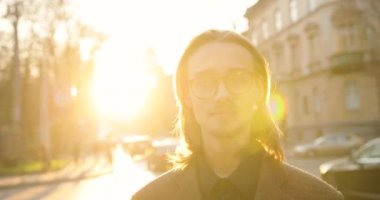 Sakallı, bıyıklı, gözlüklü, ceketli, gülümseyen ve dik bakan çekici bir genç adam portresi. Caddenin arkasında, insanlar yürüyor, arabalar sürüyor, güneş ve güneş ışığı. Güneş.
