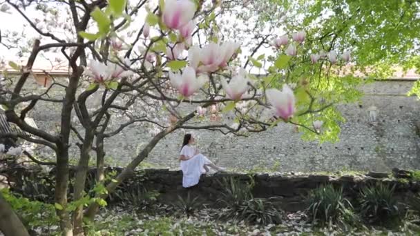 Menina morena em um fundo de magnolias rosa branco no jardim. Jovem mulher atraente em um vestido branco em um belo jardim magnólia — Vídeo de Stock
