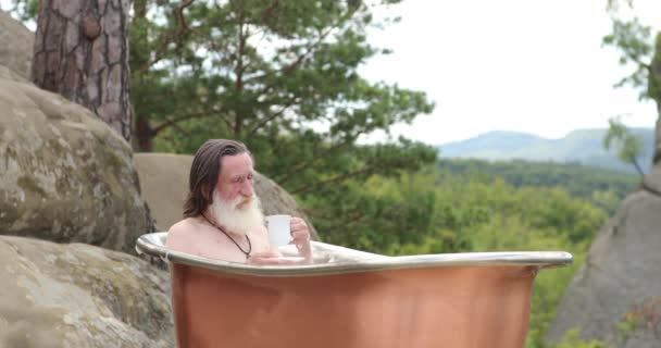 Egy öreg ember, mint a Mikulás ül és pihen a fürdőszobában és iszik egy fehér pohárból. — Stock videók